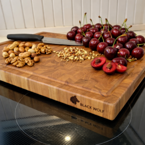 Cherry end‑grain cutting board by Black Wolf Industries with engraved logo, displayed on a black glass stovetop. The board is topped with fresh red cherries, assorted nuts, and a chef’s knife resting diagonally.