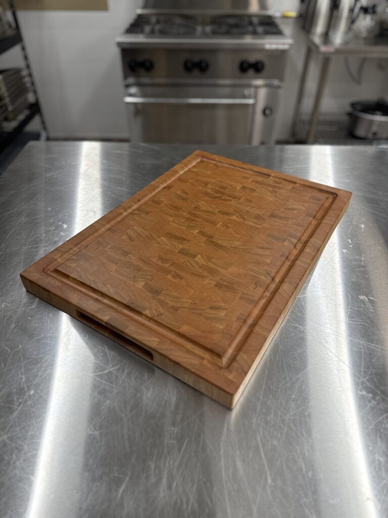 Cherry end grain cutting board sitting on a stainless steel counter in a commercial kitchen setting.